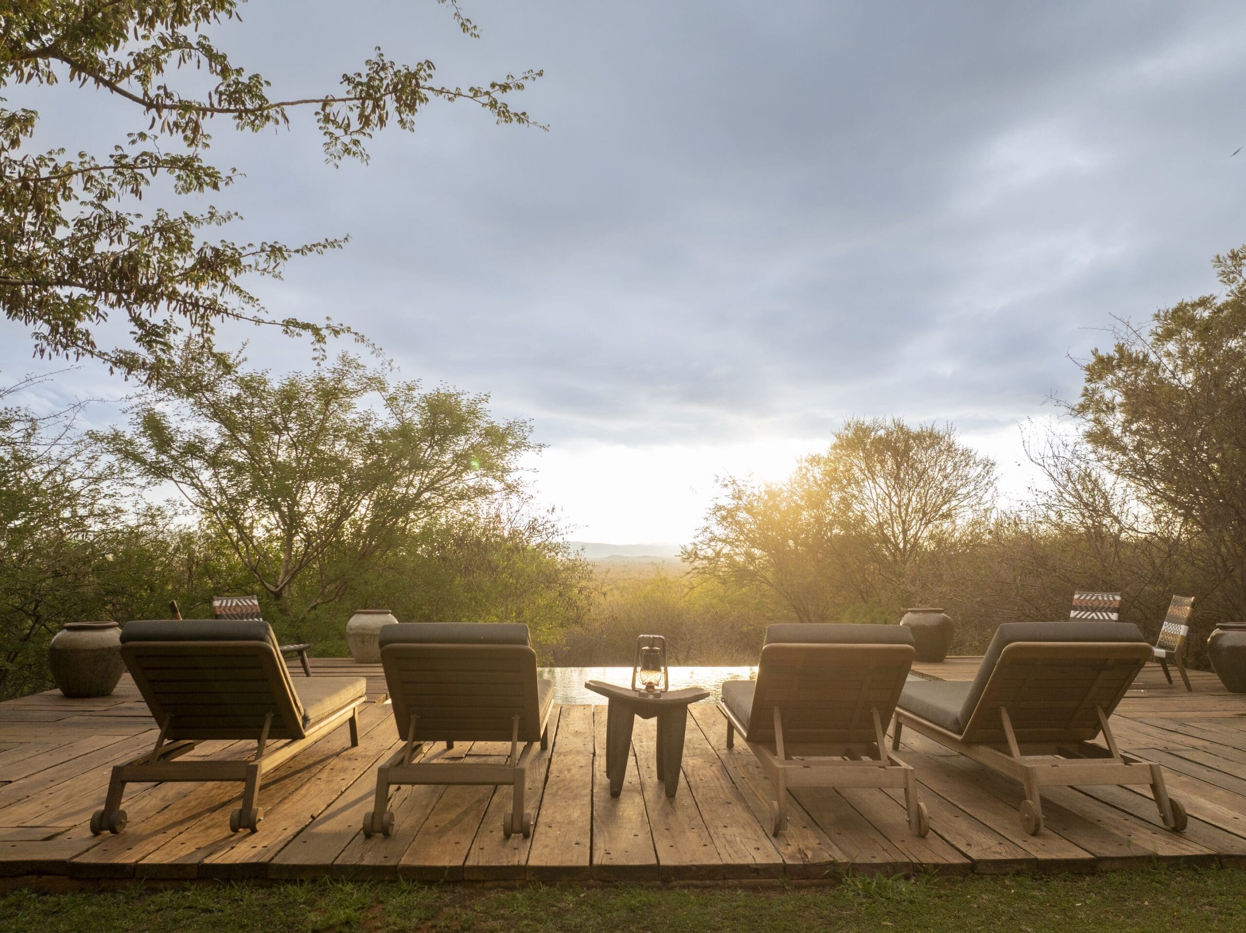Four wooden loungers face an infinity pool on a timber deck at sunset, with a lit lantern on a carved stool between them.