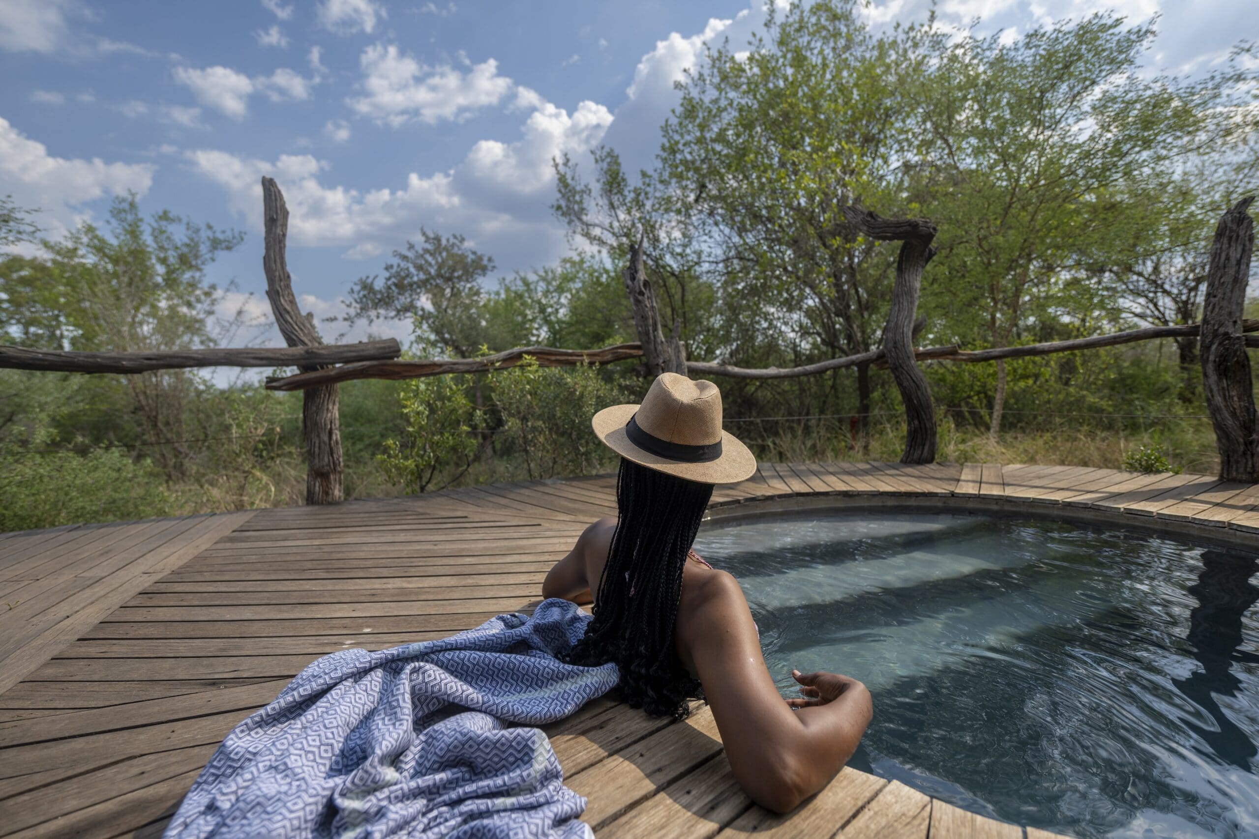 A guest in a wide-brimmed straw hat rests at the edge of a plunge pool on a timber deck, surrounded by bush.